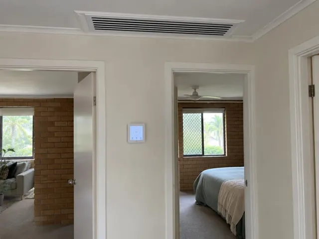 Ceiling ducted air outlet and wall controller in a cream hallway opening to brick rooms in Elanora