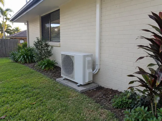 Outdoor air conditioner unit on a concrete pad beside a brick house and garden in Elanora